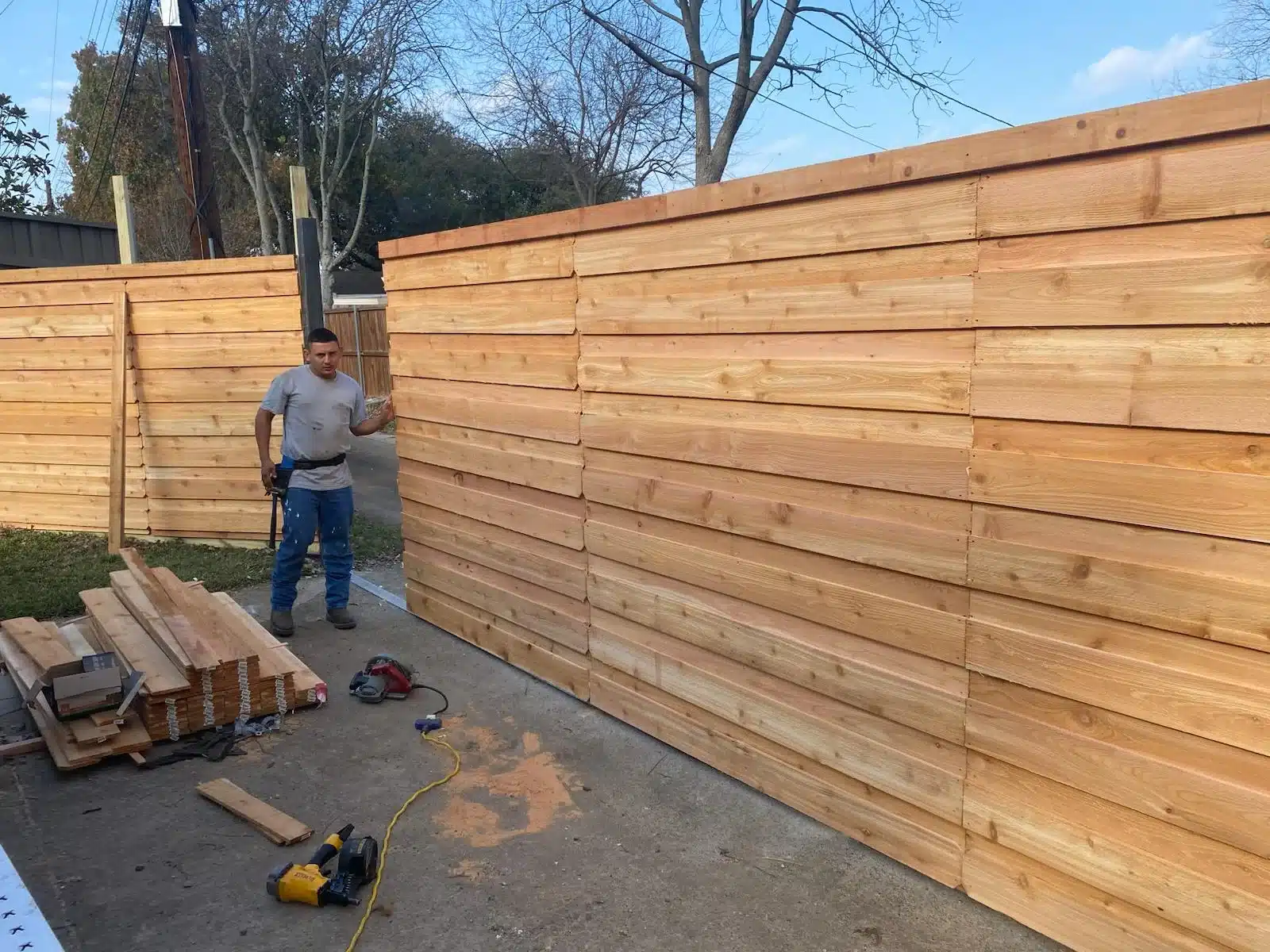 Worker beside horizontal wood panels with stacked boards, sawdust, and tools on the concrete