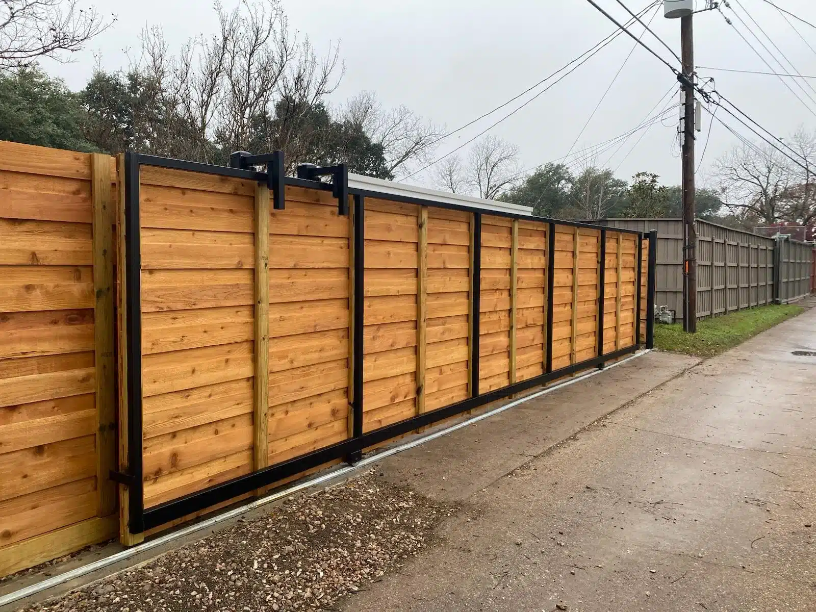 Driveway-side view of horizontal wood sliding gate with black framing, upper guides, and ground track
