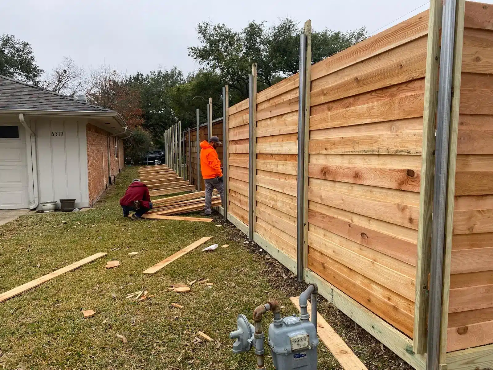Two workers beside a horizontal wood fence with metal posts, loose boards, and offcuts near a brick house.