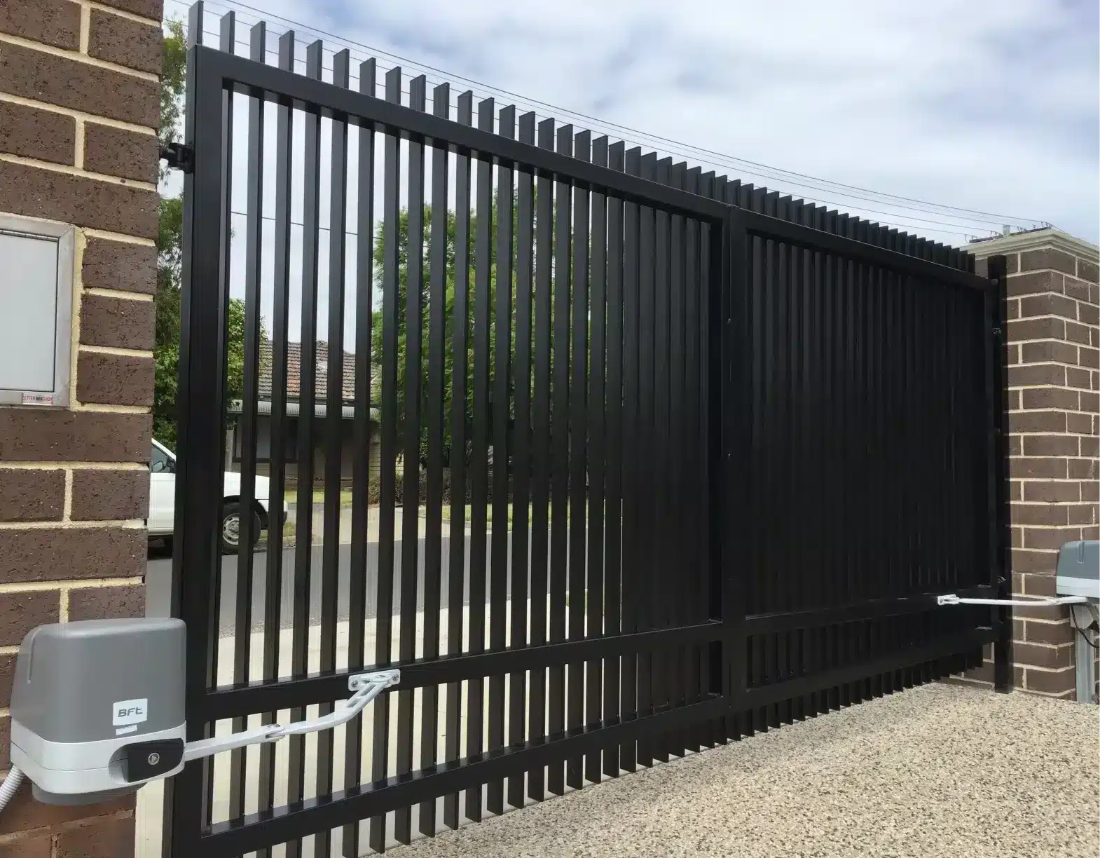 Installed black vertical-slat driveway gate between brick pillars with a mounted automated opener.