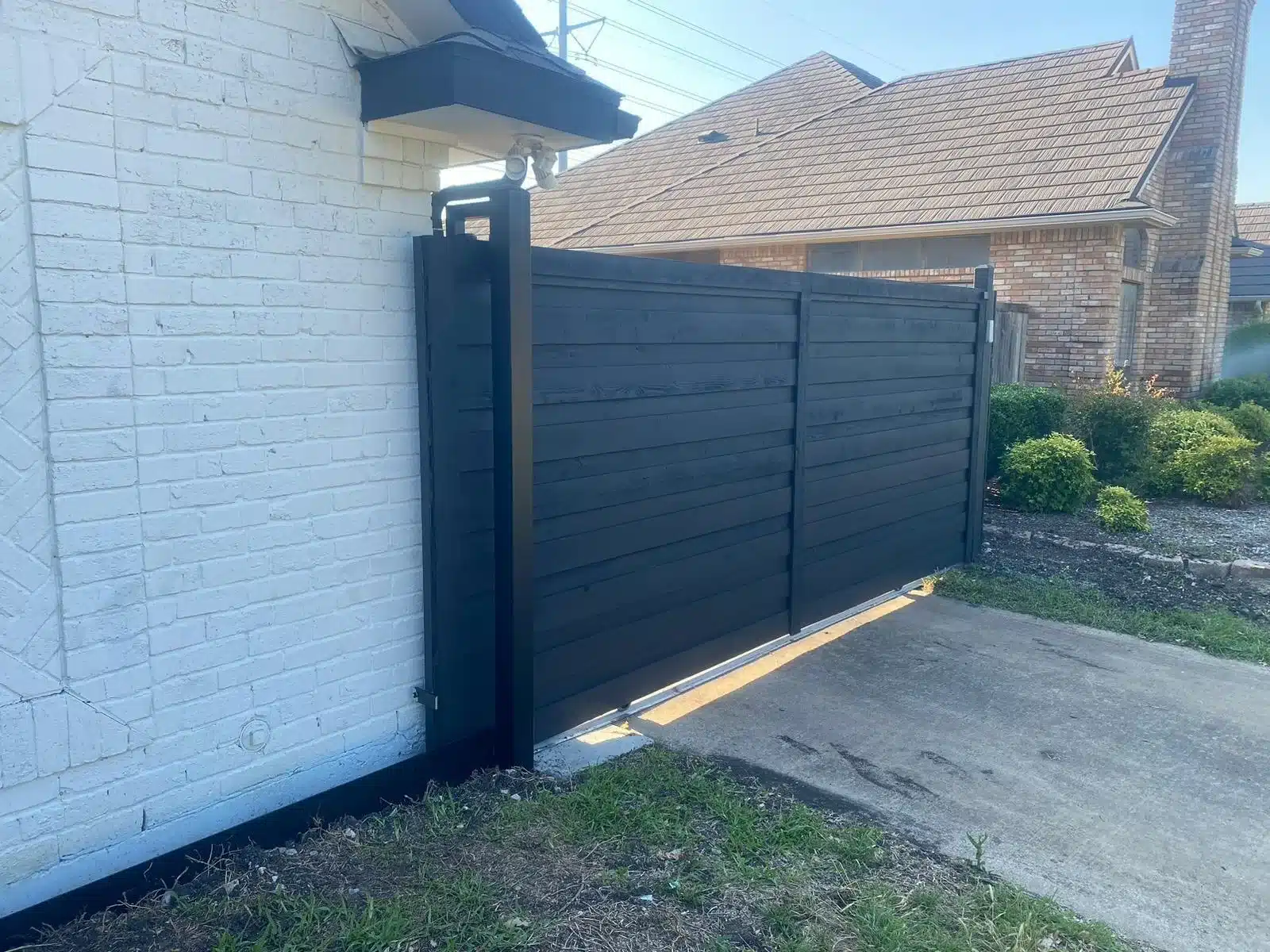 Installed black horizontal-slat driveway gate mounted on a black post beside a white brick house.