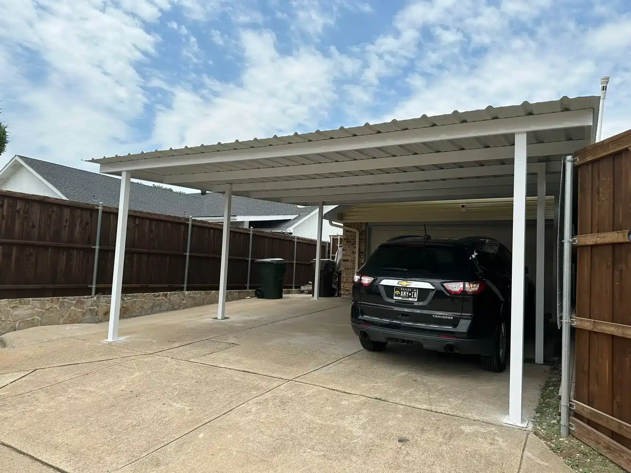 White aluminum patio cover sheltering a black Chevrolet parked on concrete driveway with dark wood fencing nearby