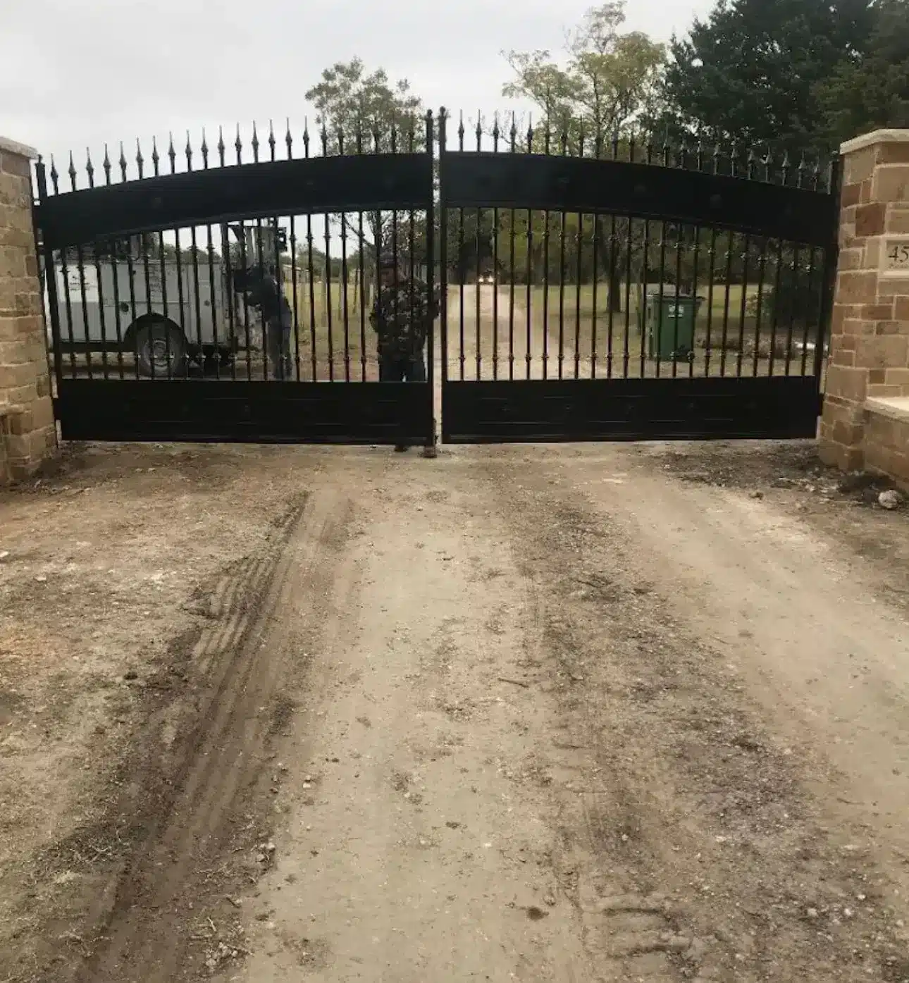 Black metal driveway gates with spear tops on brick pillars, unpaved gravel driveway beyond
