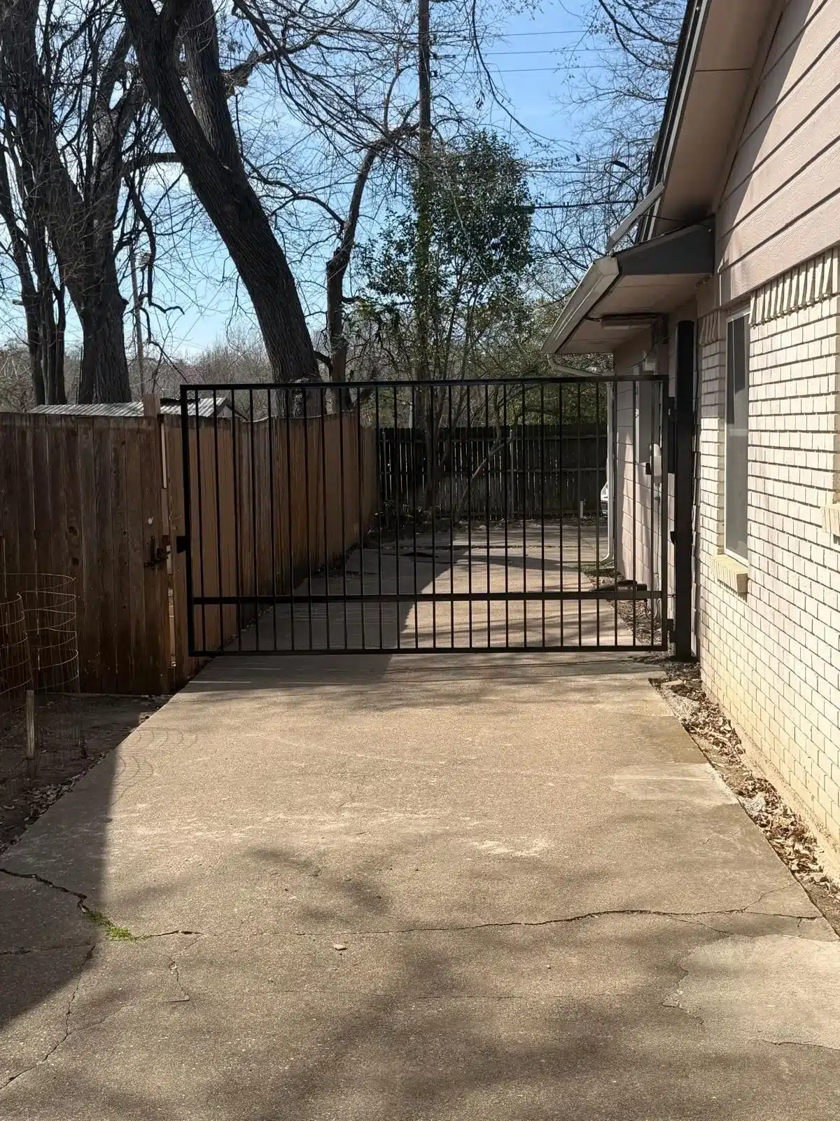 Black metal driveway gate open on concrete patio next to tan brick house with bare trees behind
