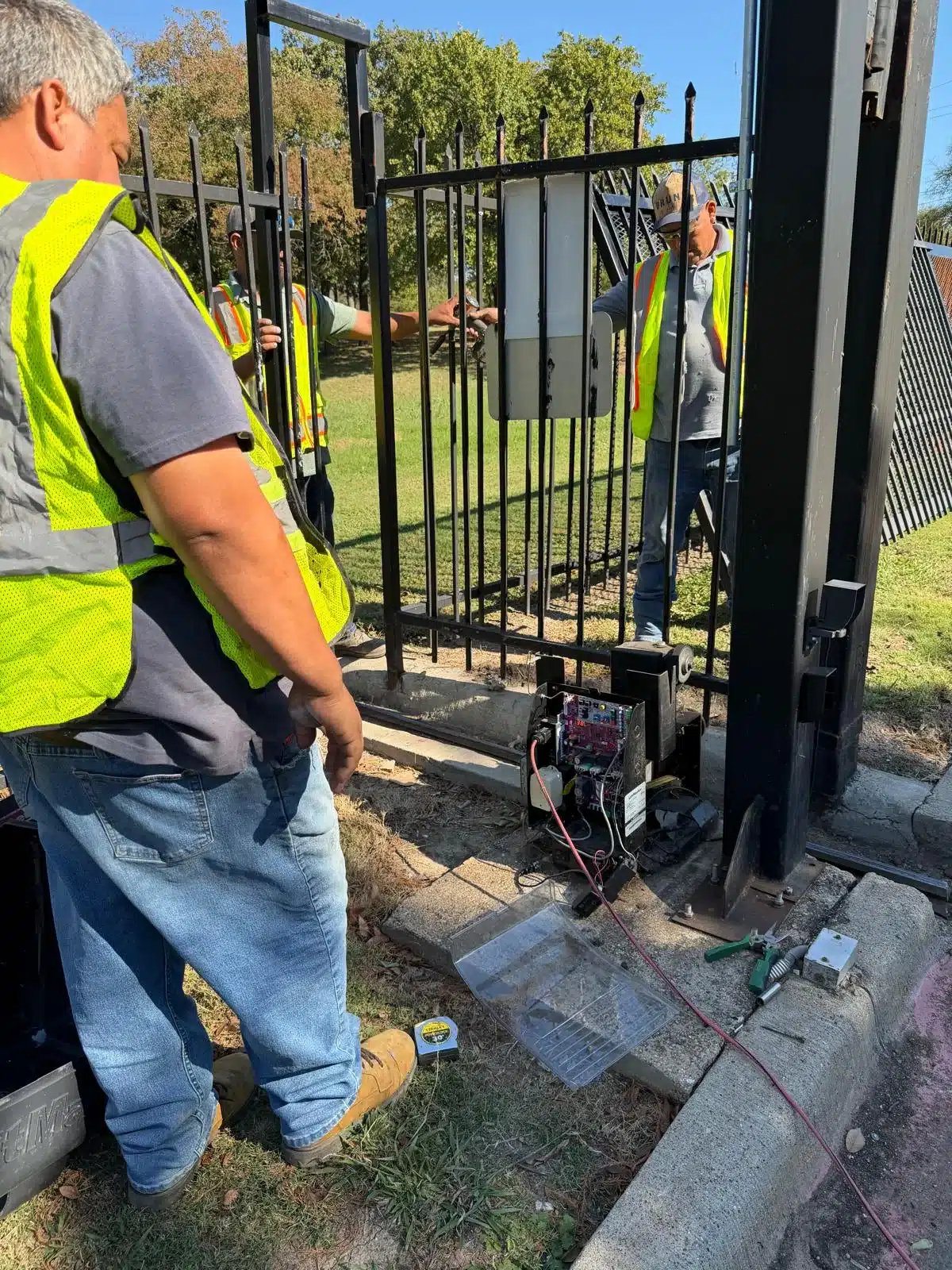 Workers installing black metal automated gate with electronic control system components.