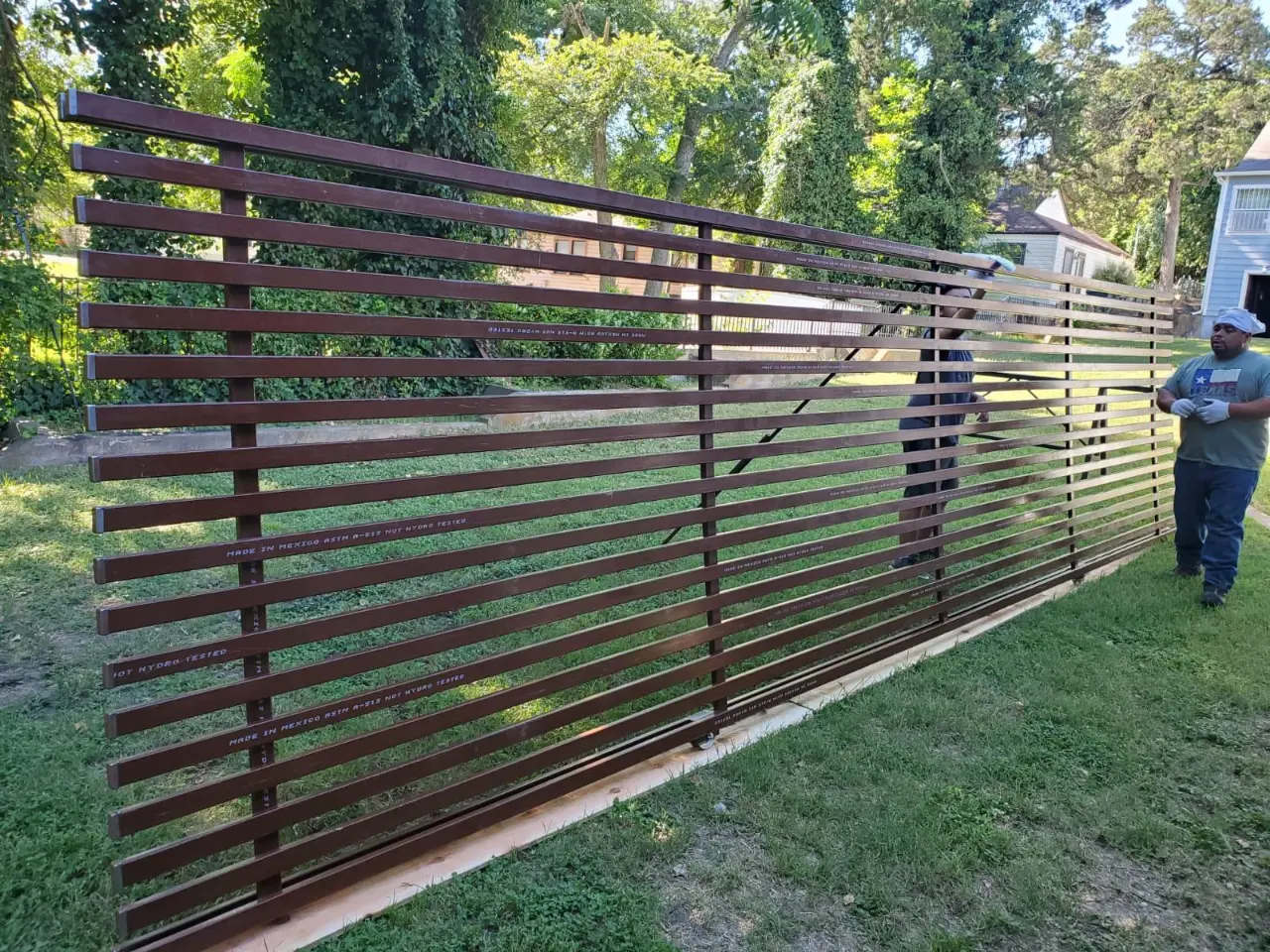 Workers installing a horizontal aluminum gate panel in a residential yard with mature trees.