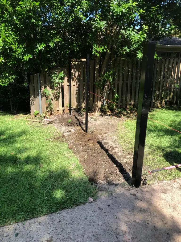 Fresh trenching line dug through a backyard with metal posts and string guides prepared for fence installation.