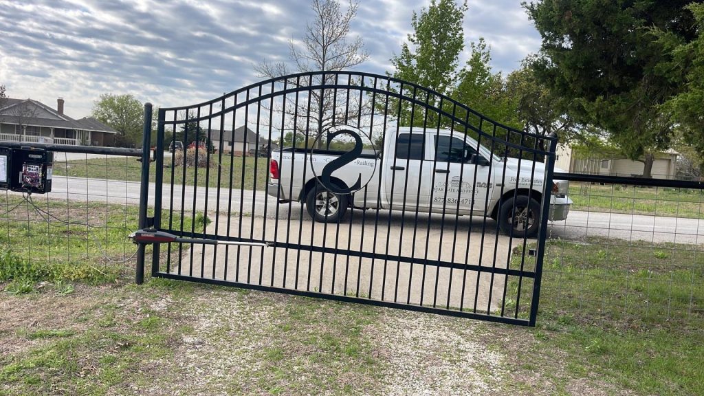 Black automated driveway gate with decorative “S” design and swing gate opener in a rural setting.