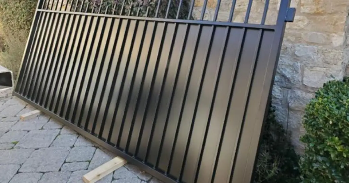 Black metal driveway gate panel resting on wooden blocks beside a stone wall, related to gates and openers.