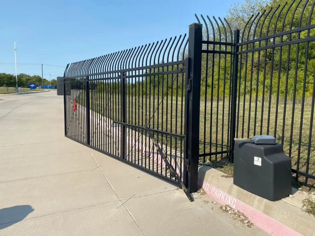 Tall black metal security gate with curved pickets and an automatic opener beside a roadway.
