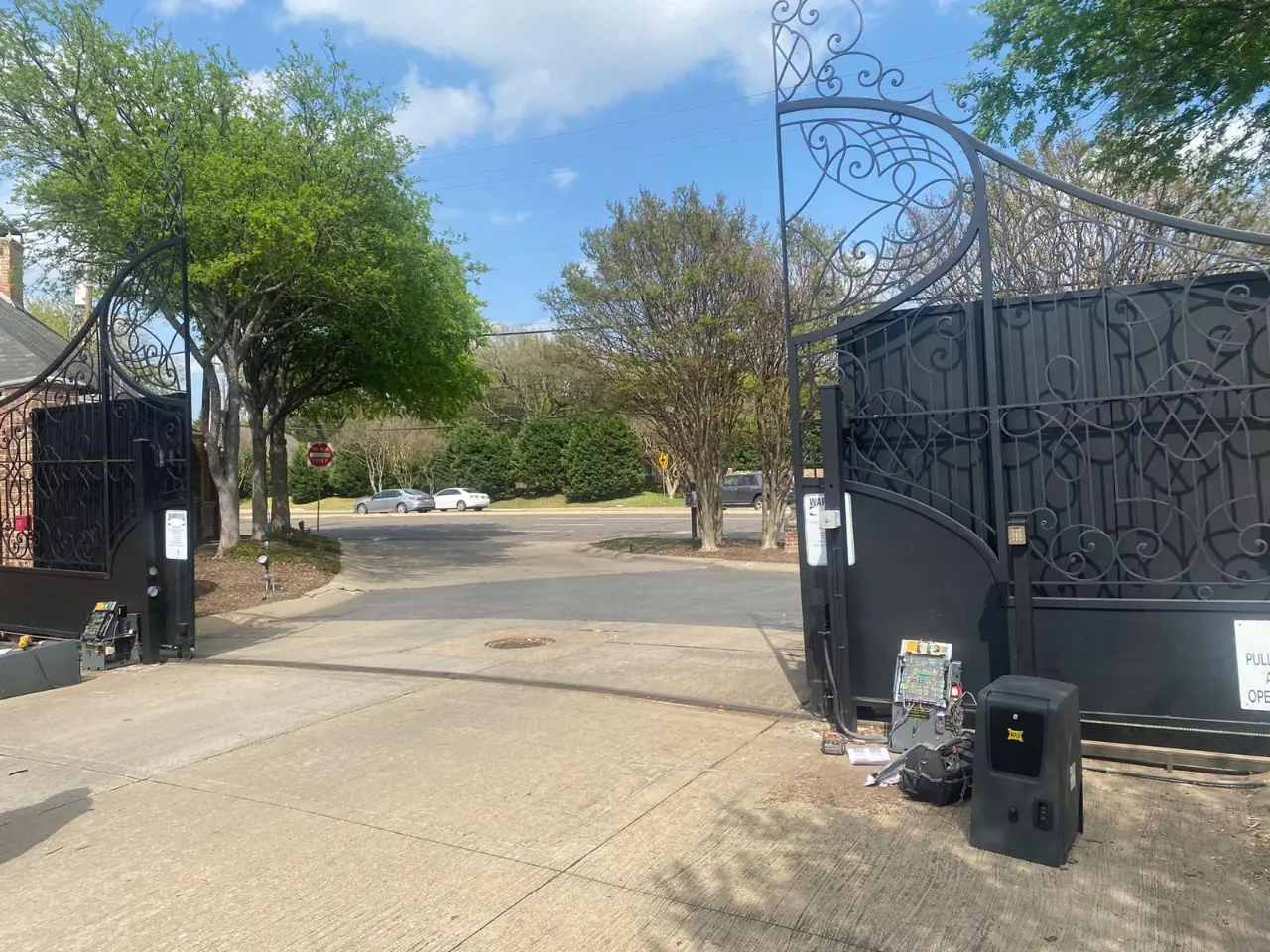 Ornate black iron gates opening onto a residential concrete driveway, with access control equipment mounted nearby.
