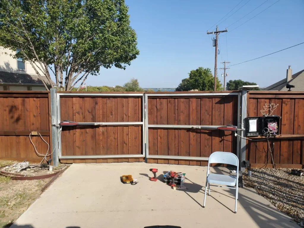 Automated wooden swing gate installed at the end of a concrete driveway, with gate motors and tools visible during installation.
