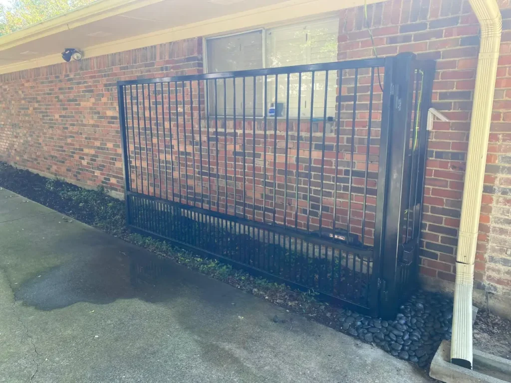 Black metal sliding gate positioned along a brick wall next to a concrete driveway and gutter downspout.