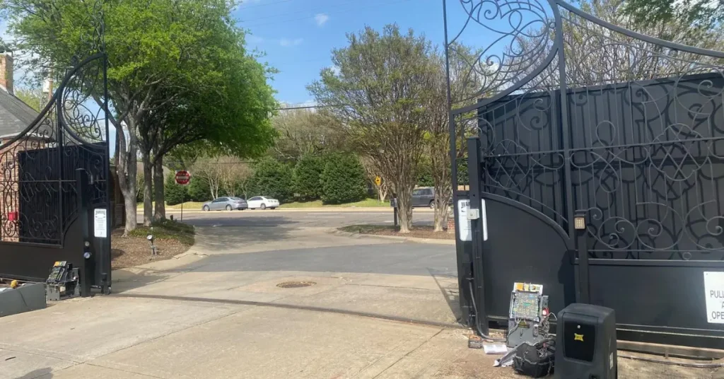 Technician repairing an ornate black driveway gate with tools and control panel open near a residential entrance.