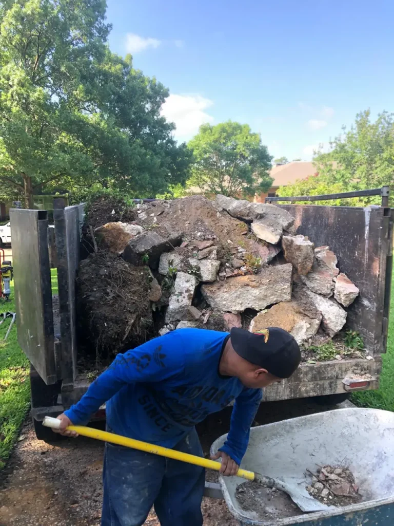 Worker shovels dirt and broken concrete into a wheelbarrow during trenching and excavation cleanup at a residential site.