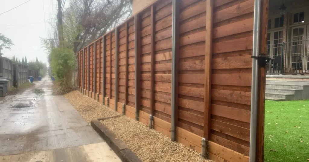 Tall horizontal wood privacy fence with steel posts lining a residential alley on a cloudy day.